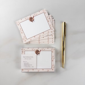 May include: A stack of rectangular notelets and a boxed set of 30 mini cards featuring a brown turkey illustration and wheat stalks. The packaging text reads NOTELETS 30 mini cards. A gold-toned metal pen is positioned to the right of the cards.