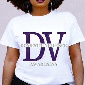 以下が含まれることがあります： 白いTシャツに紫色の「DV」のグラフィックがプリントされています。文字はグレーで縁取られており、「Domestic Violence Awareness」というテキストが文字の下に印刷されています。