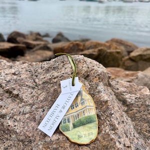 May include: An oyster shell ornament featuring a hand-painted yellow house with green landscaping, gold-painted edges, and a green ribbon. Attached is a white tag printed with "TABLE NO. 3" and "TAYLOR & ALISON HAMILTON" with "June 20, 2026" on the shell.