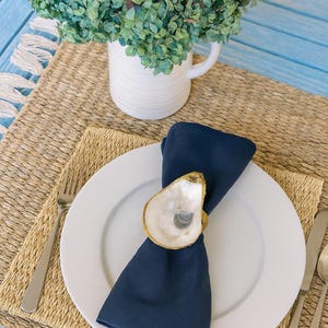 May include: A white plate with a navy blue napkin held by an oyster shell napkin ring with a gold rim. A fork and knife are on either side of the plate. A woven placemat and a white pitcher with green foliage are in the background.