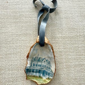 May include: An oyster shell ornament featuring a watercolor painting of a large, blue and white house. The shell has a gold-colored edge and is suspended by a gray velvet ribbon tied in a bow.