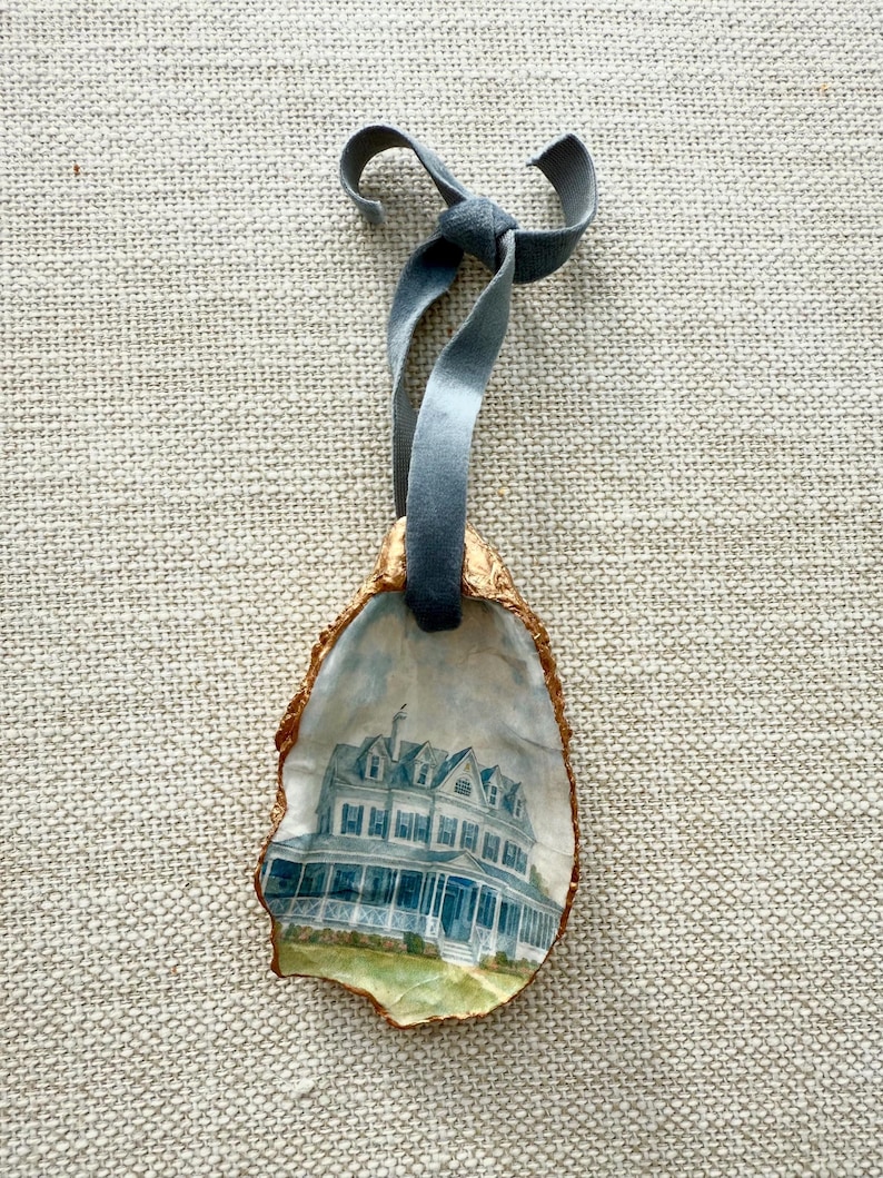 May include: An oyster shell ornament featuring a watercolor painting of a large, blue and white house. The shell has a gold-colored edge and is suspended by a gray velvet ribbon tied in a bow.