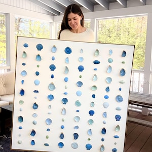 May include: A large square wall art piece featuring a white background arranged with numerous small seashells in various shades of blue, light green, and white. The shells are organized in vertical columns within a thin, metallic gold frame.