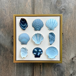 May include: A framed artwork featuring nine painted seashells in shades of blue, arranged on a white canvas. The shells have various patterns and textures, with gold accents. The frame is gold.
