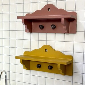 Puede incluir: Dos estantes decorativos de madera, uno rojo-marrón y otro amarillo mostaza, con bordes festoneados y dos pomos negros cada uno. Están montados en una pared de azulejos blancos. Ideales para decoración.