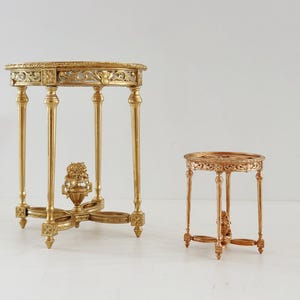 May include: Two ornate, gold-coloured side tables. The larger table has a round top with intricate carvings, supported by four slender legs and a decorative base. The smaller table mirrors the design. The tables are approximately 71 cm and 36 cm tall.