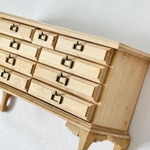 Puede incluir: Una cómoda de madera en miniatura con múltiples cajones y tiradores de color latón. La cómoda tiene un acabado de madera clara y está diseñada con patas decorativas. Esta pequeña pieza de mobiliario es probablemente para una casa de muñecas o una colección en miniatura.