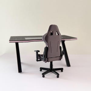 May include: Miniature gaming desk and chair set. The desk has a black surface with a pink trim and black legs. The chair is gray with black armrests and a black base. The set is designed for a miniature gaming setup.