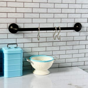 May include: A black metal bar with three silver hooks mounted on a white subway tile wall. A white colander with a blue rim sits on a white countertop below the bar.