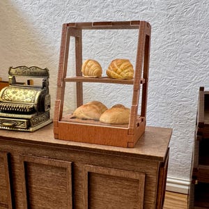 Op de afbeelding: Een miniatuur houten bakkerijvitrine met twee planken met vier kleine broodjes, geplaatst naast een koperkleurige kassa in vintage-stijl met de tekst "Amount Purchased" zichtbaar op de bovenste metalen plaat van de kassa.