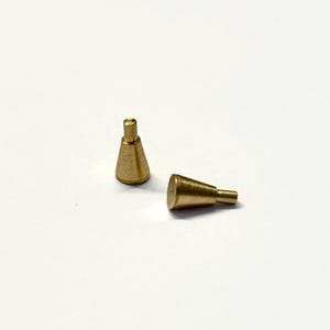 Puede incluir: Dos pequeños objetos en forma de cono, de color latón, con un vástago cilíndrico. Un objeto está de pie, mientras que el otro está tumbado de lado. Los objetos están sobre una superficie blanca.