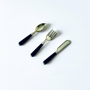 Könnte beinhalten: Ein Set aus Miniatur-Besteck, bestehend aus einem Löffel, einer Gabel und einem Messer. Jedes Teil hat einen silberfarbenen Kopf und einen schwarzen Griff. Das Besteck ist diagonal vor einem weißen Hintergrund angeordnet.