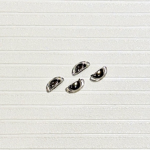 May include: Four small, semi-circular metal drawer pulls with a polished silver-tone finish and a smooth, dark black inlay. Each piece features a curved handle design with a flat base, arranged in a cluster on a textured, horizontally lined surface.