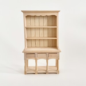 Peut inclure: Une petite armoire en bois avec trois étagères et trois petits tiroirs. L'armoire a un dessus festonné et est en bois non fini. La pièce est conçue pour une maison de poupée ou un cadre miniature.