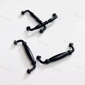 以下が含まれることがあります： 黒い金属製のキャビネットドアのプルが2つ。1つのプルはシンプルなデザインで、もう1つはねじれたディテールが施されたより装飾的なデザインです。