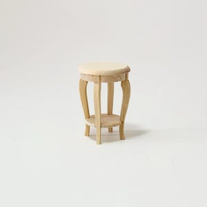 May include: A small, unfinished wooden side table with a round top and curved legs. The table has a lower shelf and is designed for decorative purposes. The natural wood color gives it a rustic, vintage feel. The table is approximately 12 inches tall.