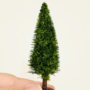 May include: A miniature model evergreen tree featuring a conical shape with textured dark green foliage and a brown trunk. The small scale tree has a pointed top and a flat base, designed for use in dioramas, model train layouts, or craft projects.