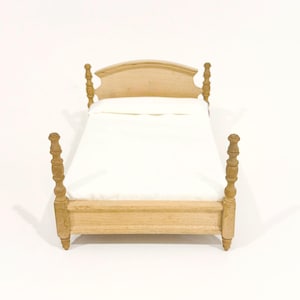 May include: A miniature wooden bed frame with a light tan finish, featuring four turned posts and a curved headboard. The bed is dressed with a plain white mattress and a matching white pillow, creating a simple and clean appearance for the furniture.