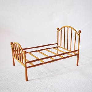 Puede incluir: Un pequeño marco de cama de madera con un acabado marrón claro. La cama tiene cabecero y piecero con listones verticales y remates redondeados. El marco tiene un diseño simple y minimalista, adecuado para una muñeca o miniatura.