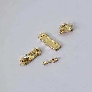 Könnte beinhalten: Vier kleine, goldfarbene dekorative Hardware-Teile. Enthält einen Schlüssel, eine rechteckige Platte, ein dekoratives Stück mit einem klaren Edelstein und ein kleines, verziertes Stück. Wahrscheinlich für die Schmuckherstellung oder Bastelprojekte.