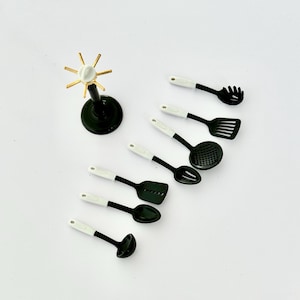 May include: A set of miniature black and white kitchen utensils, including a ladle, slotted spoon, spatula, and whisk. A dark green stand with a gold and white starburst design is also present. The utensils are arranged on a white surface.
