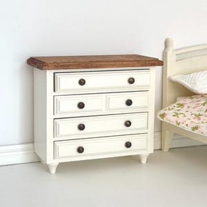 Peut inclure: Une commode miniature en bois avec un plateau teinté brun foncé et une structure peinte en blanc. Le meuble comprend quatre tiroirs dotés de petits boutons ronds foncés. Le tiroir supérieur est pleine largeur, tandis que le second a deux tiroirs.