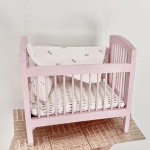 May include: A miniature pink cot with vertical bars and a curved headboard. The cot is furnished with a patterned mattress, pillow, and a blanket with bow tie designs. The cot is placed on a patterned surface, suggesting a dollhouse or nursery theme.