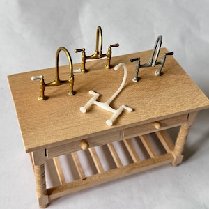 Puede incluir: Cuatro grifos de cocina en miniatura en diferentes acabados, incluyendo oro, plata y blanco, se muestran en una encimera de madera. Los grifos están dispuestos en una fila, con el grifo de oro a la izquierda, el grifo de plata en el medio y el grifo blanco a la derecha. La encimera es parte de una mesa de madera con un estante debajo.