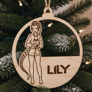 May include: Wooden Christmas ornament featuring a cutout of a woman holding a gift, with the name "LILY" below. The ornament is hanging on a Christmas tree. The woman has a long braid.