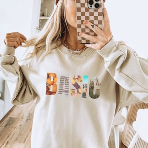 Könnte beinhalten: Ein beiger Pullover mit einem grafischen Aufdruck des Wortes "BEACH" in bunten, gemusterten Buchstaben. Das Model trägt den Pullover mit braunen Shorts und weißen Sneakern.