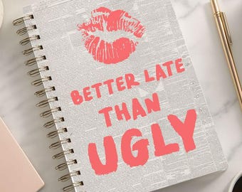 Puede incluir: Un cuaderno con espiral con fondo gris y el texto "BETTER LATE THAN UGLY" en coral. Una huella de labios coral está encima del texto. Un bolígrafo dorado, una taza de café y macarons también están en la superficie de mármol.