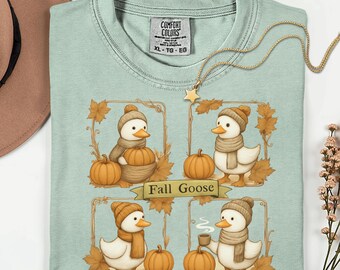 May include: A light blue Comfort Colors t-shirt featuring a "Fall Goose" design. The design includes four panels with cartoon geese wearing hats and scarves, pumpkins, and fall foliage. A gold necklace with a star pendant is draped over the shirt.