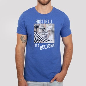 Pu&ograve; includere: Una t-shirt blu reale con una stampa grafica bianca di due giocatori di hockey e un arbitro. Il testo "FIRST OF ALL I'M A DELIGHT" &egrave; stampato in bianco sotto l'immagine.