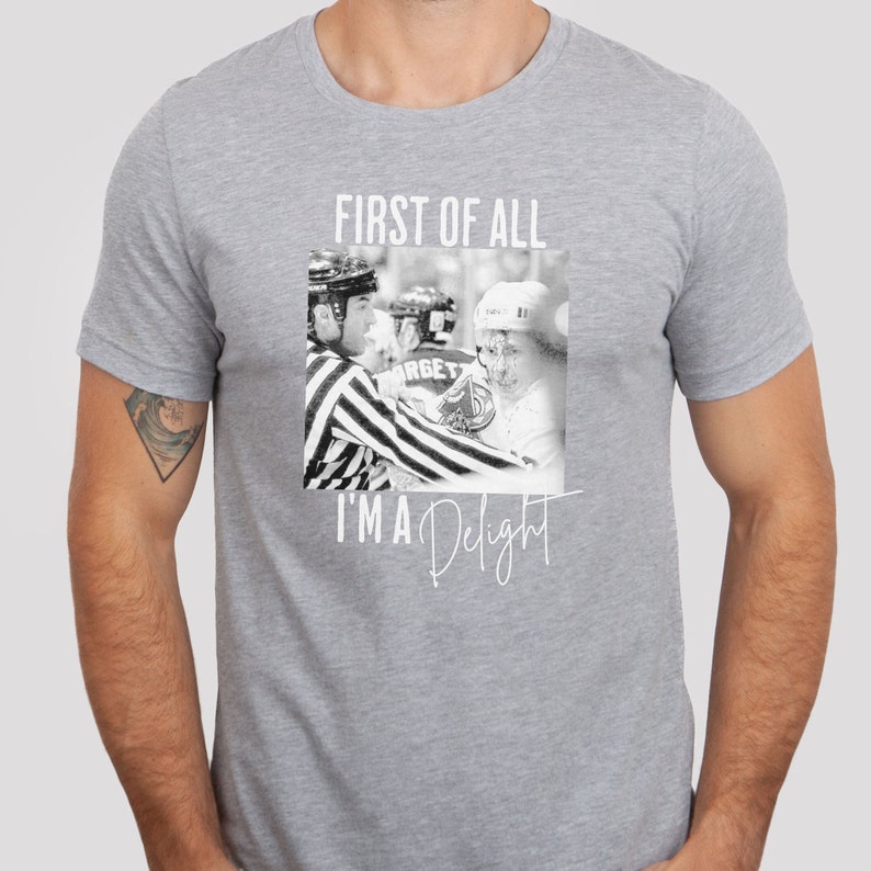 Może przedstawiać: Szary T-shirt z czarno-białym zdjęciem dw&oacute;ch hokeist&oacute;w. Na koszulce nadrukowany jest tekst "FIRST OF ALL I'M A DELIGHT".
