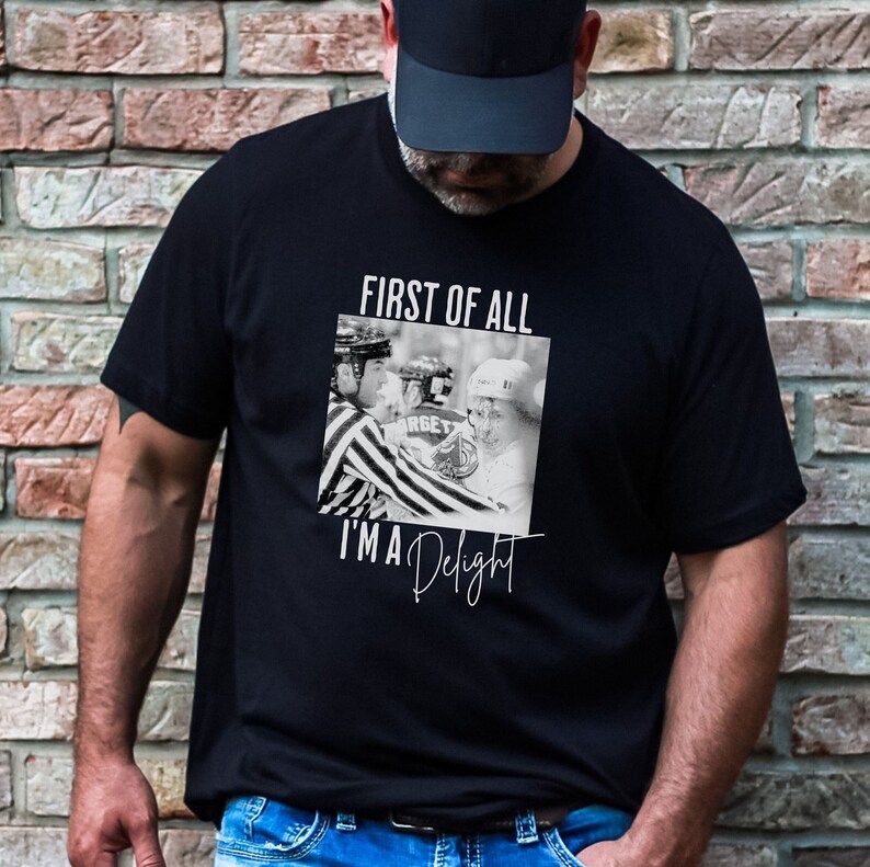 A man wearing a black tshirt with a photo image of a hockey player with blood running down his face, looking angry, being held back by a referee. The shirt reads First of All I'm a Delight.
