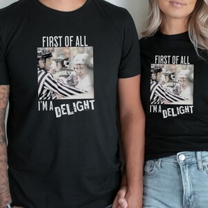 Pu&ograve; includere: Due magliette nere con una grafica di un giocatore di hockey e il testo "First of all, I'm a delight".