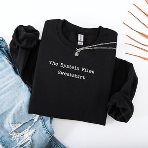 Może przedstawiać: Czarna bluza z białym napisem "The Epstein Files Sweatshirt". Srebrny naszyjnik z okrągłą zawieszką leży na ubraniu. Bluza jest prezentowana z niebieskimi dżinsami na białej powierzchni.