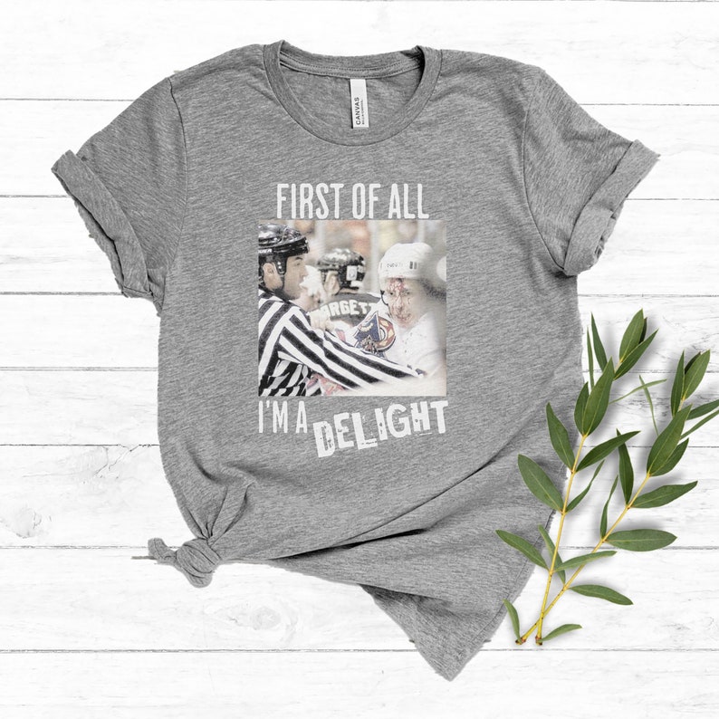 Pu&ograve; includere: Maglietta grigia con il testo "First of all I'm a delight" e una foto di un giocatore di hockey che riceve una penalit&agrave;.
