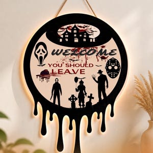 Op de afbeelding: Een rond wandbord met een zwarte rand en een druipend verfeffect. Het midden toont een spookhuis, silhouetten van horrorfilmfiguren en de tekst "WELCOME YOU SHOULD LEAVE" in zwarte en rode letters op een crèmekleurige achtergrond.