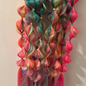 May include: Two sets of colorful, rainbow-hued hair extensions. Each set features multiple bulbous sections of vibrant, twisted hair, secured with small bands. The ends of the extensions are long and flowing, with a bright pink color.