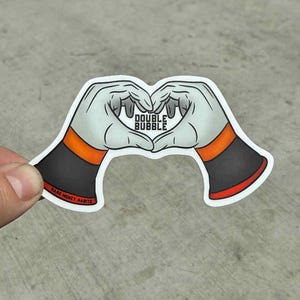 May include: A sticker featuring two cartoon hands forming a heart shape. The hands are white with black and orange wristbands. The text "DOUBLE BUBBLE" is in the center of the heart. The sticker has a white border.