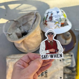 May include: A white sticker with a cartoon illustration of a man wearing a white hard hat, sunglasses, and a red vest. The text "SAY/WHEN" is printed below the illustration.
