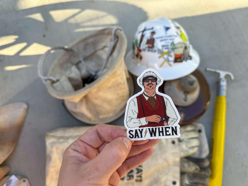 May include: A white sticker with a cartoon illustration of a man wearing a white hard hat, sunglasses, and a red vest. The text "SAY/WHEN" is printed below the illustration.