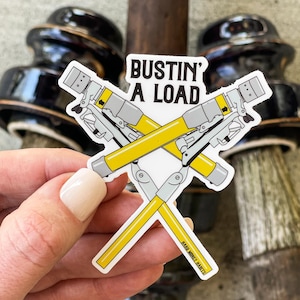 May include: A white sticker with the text "BUSTIN' A LOAD" and a graphic of two crossed yellow and gray tools. The sticker also has the text "HARD MONEY HABITS" in the bottom right corner.