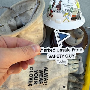 May include: A white hard hat with various safety stickers, including "Marked Unsafe From Safety Guy Today" and "Always Wear Your Gloves". The hard hat is next to a yellow tool and a white bucket.