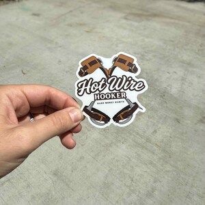 May include: A white sticker with the text "Hot Wire Hooker" and "Hard Money Habits" in a stylized font. The sticker features a graphic of two brown and tan tools. The sticker is held by a hand.