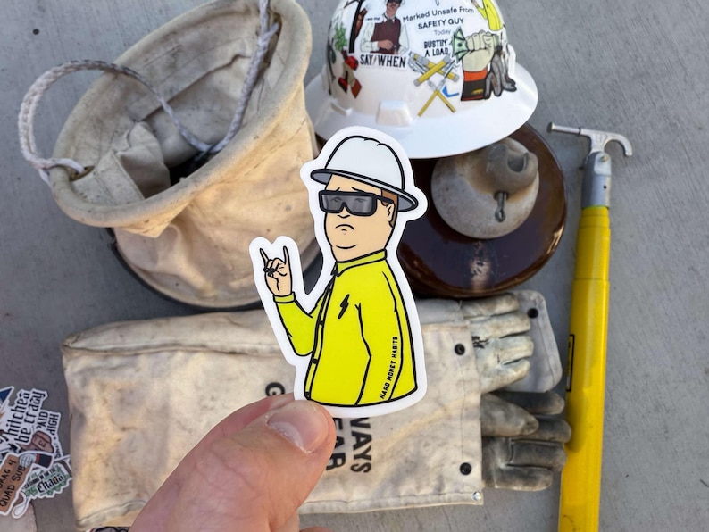 May include: A sticker of a cartoon character wearing a yellow jacket and a hard hat. The character is giving a rock and roll hand gesture. The sticker is on a white background and has the text "Hard Money Marts" on the bottom right corner.