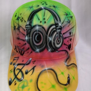 May include: A baseball cap with an airbrushed design. The cap features a large pair of headphones, starbursts, and musical notes. The brim has the text "XOXO" and a musical staff. The colour scheme includes green, red, and yellow.