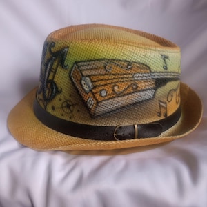 May include: A straw fedora hat with a black leather band and buckle. The hat is painted with a musical instrument and musical notes in shades of yellow, orange, and brown. The brim is a light tan colour.