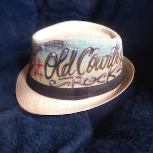 May include: A beige fedora hat featuring a black band and the text "Old Country Rock" on the crown. The hat has a light-coloured brim and a colourful design on the crown.